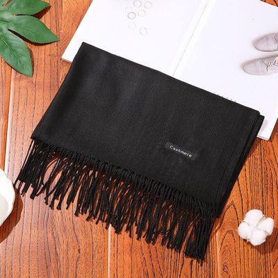 Black cashmere women's scarf shawl with fringe on wooden surface near decor items