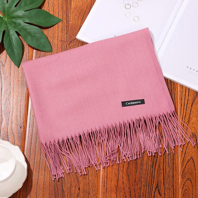 Pink cashmere women scarf shawl with fringes on wooden surface beside green leaf and white accessories
